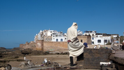 Man in a bernous in front of the old city walls and buildings inside the medina in Essaouira,...
