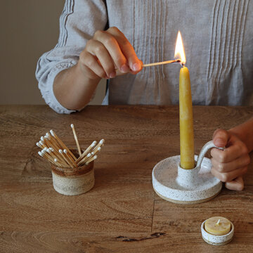 Female seated at wooden table lighting a beeswax candle. 