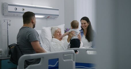 Female elderly patient lies in bed in bright hospital ward, plays with little grandson. Loving...
