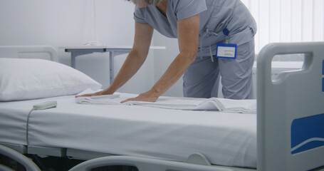 Mature cleaner changes bedclothes and puts clean pillow on bed. Nurse makes beds in hospital ward...
