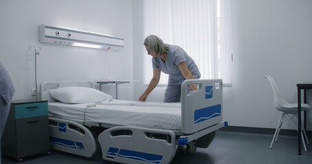 Mature cleaner changes bedclothes and puts clean pillow on bed. Nurse makes beds in hospital ward and prepares it for new patients in bright hospital ward. Medical staff work in modern medical center.