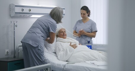 Obraz premium Mature nurse sets up bed using remote control, prepares hospital room for patient. Female medical worker pushes wheelchair with elderly woman and puts her in bed. Modern clinic or medical facility.