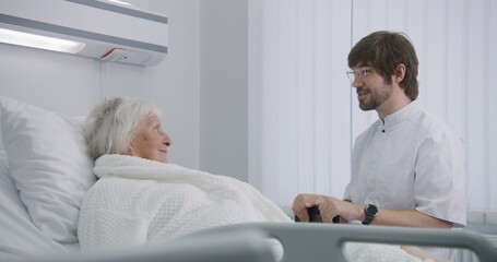 Obraz premium Doctor consults female elderly patient lying and resting in bed in hospital room. Male physician uses digital tablet, talks with nurse. Senior woman recovering after successful surgery. Modern clinic.