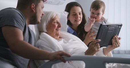 Hospital room. Old woman lying in bed spends time with relatives. Loving family use tablet...