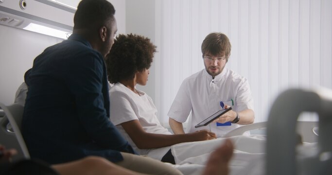 Physician reports test results to clients using tablet computer. African American boy and his father consults with doctor. Patient recovering after surgery. Medical personnel work in hospital ward.