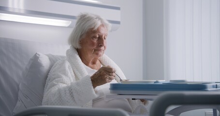 Elderly woman eats delicious food and looks at camera resting in bed in bright hospital ward....