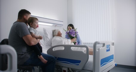 Elderly woman lying and resting in bed in bright hospital ward, plays with grandson. Loving family members support grandmother recovering after successful surgery. Modern medical facility or clinic.