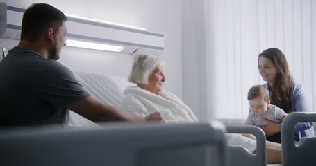 Elderly woman lying and resting in bed in bright hospital ward, plays with grandson. Loving family members support grandmother recovering after successful surgery. Modern medical facility or clinic.