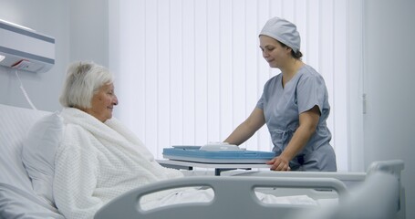 Obraz premium Nurse opens big lunch box with meal to female old patient. Elderly woman resting in bed in bright hospital room, recovering after successful surgery, eating food. Modern medical facility or clinic.