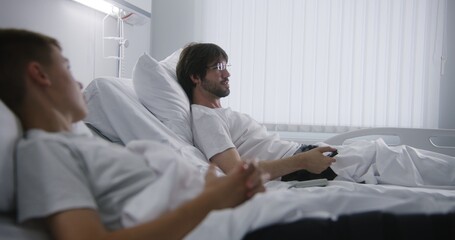 Adult man talks with kid, puts on glasses and turns on TV. Patients rest together in hospital ward. Hospital roommates fully recovering after successful operations. Modern and bright medical center.