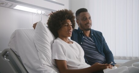 Happy African American teenager rests in hospital room and watches movie together with father....
