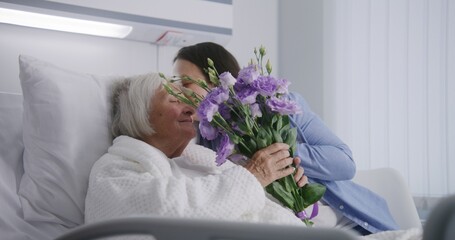 Loving family with little child visits grandmother recovering after successful surgery, gives flowers. Elderly woman lies and rests in bed in bright hospital ward. Modern medical facility or clinic.