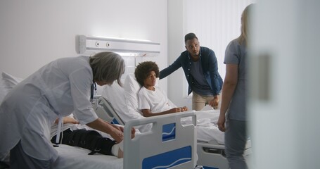 Obraz premium Mature physician bandages leg of young boy. African American man visits son in hospital ward. Nurse comes to teenager and raises back of his bed. Patient fully recovering after successful operation.