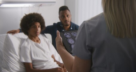 Physician reports X-Ray results to clients using tablet computer. African American teenager lies on bed and talks with doctor. Patient recovering after operation. Medical staff work in hospital ward.
