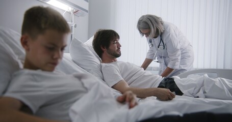 Obraz premium Man rests in bed, talks with young kid and turns on TV. Mature doctor does checkup of patients in hospital ward. Hospital roommates fully recovering after successful operations. Modern bright clinic.