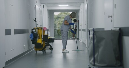 Mature cleaner mops floor in medical center corridor. Multicultural teens stand near window and talk. Caucasian boy with broken leg leaves his friend and goes to hospital ward down clinic hallway.