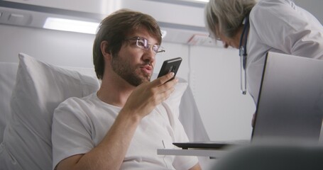 Adult man lies on bed in hospital ward and chats with client using phone. Man in glasses remotely...