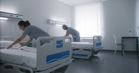 Adult nurse comes to hospital room and starts to change bedclothes with her mature colleague. Female cleaners make beds and prepare it for new patients. Medical personnel at work in modern clinic.