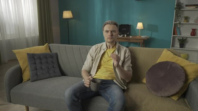 Zoom in video of a middle-aged man sitting on a couch, sofa, drinking tea, when suddenly he feels a strong toothache, pain and touches his cheeck. HDR BT2020 HLG Material.