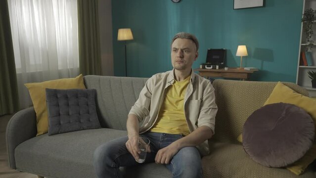 Zoom in video of a middle-aged man sitting on a couch, sofa, drinking water, when suddenly he feels a strong toothache, pain and touches his cheeck. HDR BT2020 HLG Material.