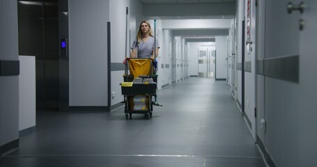 Cleaner goes from hospital room after cleaning. Health worker pushes cleaning trolley down clinic...