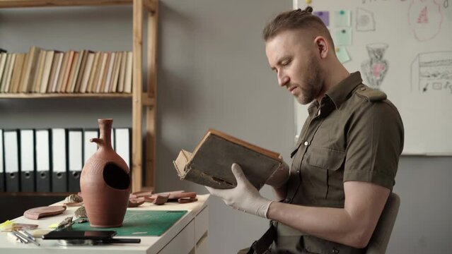 Male archaeologist working in the office studying ancient artifacts reading old book sitting at his desk.