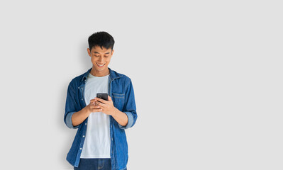 Handsome Asian man wearing a blue jeans shirt is happily playing on a mobile phone isolated on a gray background. Typing and chatting with friends via smartphone. Online social concept. Clipping path