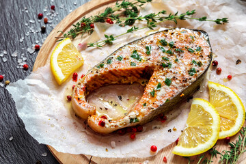 Fried salmon steak with thyme and lemon served on wooden plate on black table.