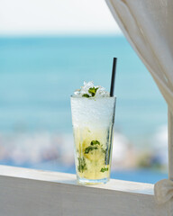 Glass of majito cocktail on terrace against blue sea. Cool drink on hot sunny day in cafe by the sea