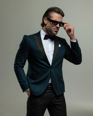 sexy young guy in tuxedo holding hand in pocket and adjusting sunglasses