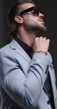 project video of stylish young man with sunglasses touching beard, looking to side, arranging suit and crossing arms and posing on grey background