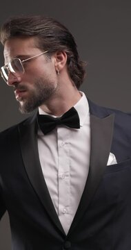 stylish elegant man wearing black tuxedo looking to side and arranging eyeglasses in front of grey background
