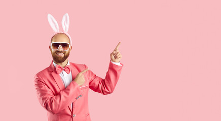 Look here. Happy cheerful handsome bearded man in pink suit, sunglasses and cute funny Easter Bunny ears standing and pointing to side at something on blank colour advertising copyspace background