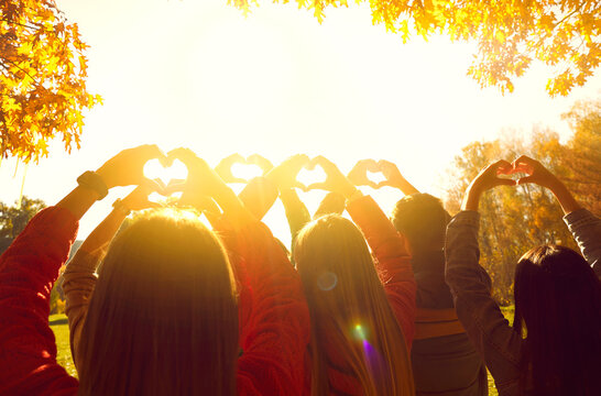 Group Of People Greeting Sunrise In Nature. Young Adult Male And Female Friends Standing In Autumn Park At Dawn, Holding Up Hands And Forming Heart Shapes In Bright Sunlight. Back View From Behind