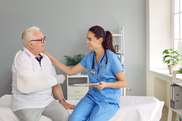 Friendly doctor supporting senior patient with broken arm. Smiling young woman in scrubs uniform...
