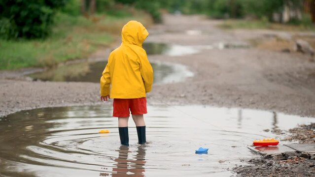 A 3-4 year old boy in a yellow waterproof raincoat and rubber boots launches boats in a puddle in the rain. Children's fun and street games