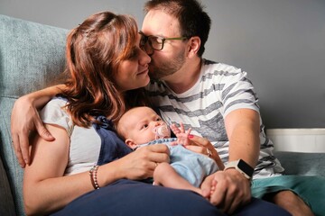 Caucasian parents kissing sitting with their one month daughter on the sofa at home. Baby girl care.