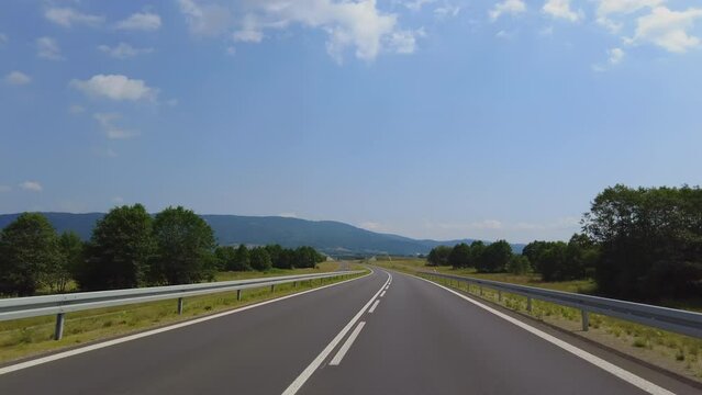 Low angle car bumper pov driving on asphalt, empty road trough mountains environment in a sunny day with blue sky and white clouds.