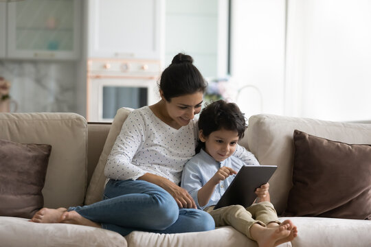 Smiling Indian mother with son using tablet at home, relaxing sitting on cozy couch together, loving young mom and 5s boy child looking at gadget screen, watching video or cartoons, playing games