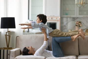 Loving laughing Indian mother lying on couch, lifting 5s son pretending flying super hero, smiling...