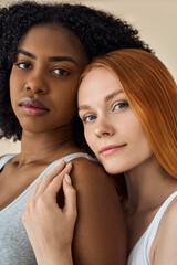 Two confident diverse women, African and Caucasian young girls friends models pretty faces bonding looking at camera standing isolated on beige background. Diversity and skin care, vertical portrait.