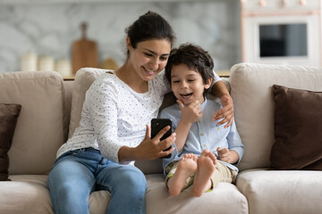 Smiling Indian mother and son having fun with smartphone together, hugging, sitting on cozy sofa,...