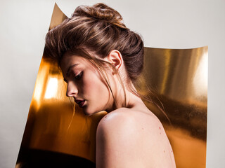 Portrait of female fashion model with naked shoulders and long hair looking down over gold background. Beautiful woman with trendy make-up