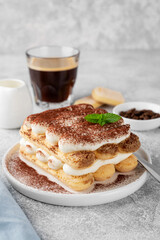 Tradition italian layered dessert tiramisu with mascarpone cream and biscuits on a white plate with cup of coffee on a gray concrete background.