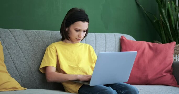 A diligent schoolboy sits on a couch, engrossed in his laptop. With an air of concentration, he types away on the keyboard, balancing schoolwork and comfort effortlessly.