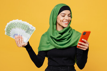 Young happy muslim woman wear green hijab abaya black clothes hold fan of dollar cash money use mobile cell phone isolated on plain yellow background People uae middle eastern islam religious concept