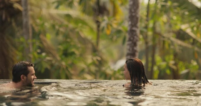 Young adult couple in love swimming together in an open-air infinity pool enjoying a tropical vacation on a villa looking away. The boyfriend and his girlfriend swimming and admiring the landscape