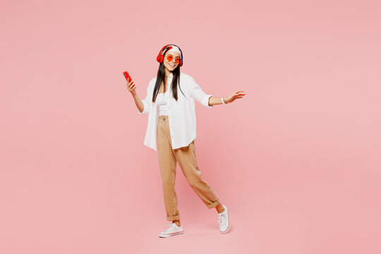 Full Body Side View Young Latin Woman She Wearing White Shirt Casual Clothes Sunglasses Dance Listen To Music In Headphones Use Mobile Cell Phone Isolated On Plain Pastel Light Pink Background Studio.