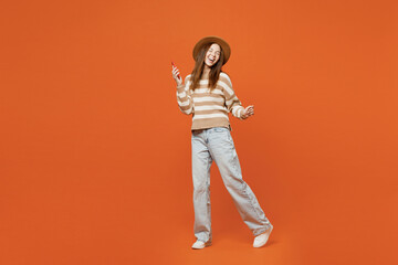 Full body side view young happy woman wear striped sweater hat casual clothes hold in hand use mobile cell phone isolated on plain orange red color wall background studio portrait. Lifestyle concept.