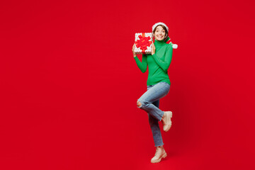 Full body overjoyed young woman wear green turtleneck Santa hat posing hold present box with gift ribbon bow isolated on plain red background Happy New Year 2024 celebration Christmas holiday concept